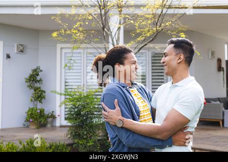 Biracial couple enjoys a warm embrace outdoors, with copy space. They share a joyful moment in a serene home garden setting. Stock Photo