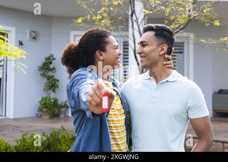 Young biracial man and Middle Eastern man smile outdoors, holding keys. They celebrate homeownership in a sunny suburban setting. Stock Photo