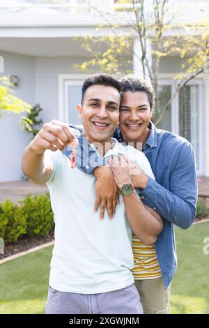 Biracial gay couple holding keys outdoors, unaltered. They celebrate homeownership with a joyful embrace in their new garden. Stock Photo