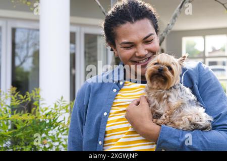 Young biracial man enjoys time with his dog outdoors, with copy space. He smiles broadly, showcasing a moment of joy and companionship in a home garde Stock Photo