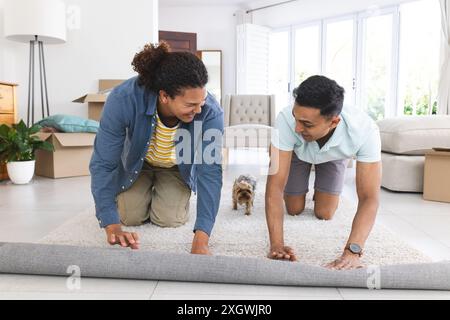 Gay biracial couple enjoys playtime with their dog at home. They're rolling out a rug, creating a cozy space for family activities. Stock Photo
