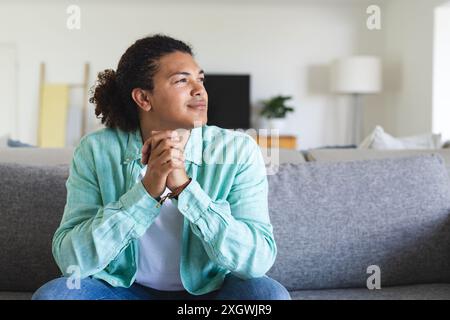 Young biracial man sits thoughtfully on a couch at home. He appears contemplative, reflecting on personal matters or decisions. Stock Photo