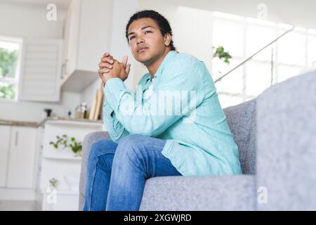Young biracial gay couple sits thoughtfully on a couch at home. They appear contemplative, reflecting on personal matters or decisions. Stock Photo
