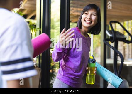 Happy asian female friends holding yoga mats and talking at home. Domestic life, friendship, activity and lifestyle, unaltered. Stock Photo