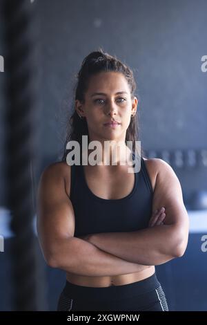 A strong and fit young biracial woman stands at the gym unaltered. Her posture exudes strength and determination in a fitness setting. Stock Photo
