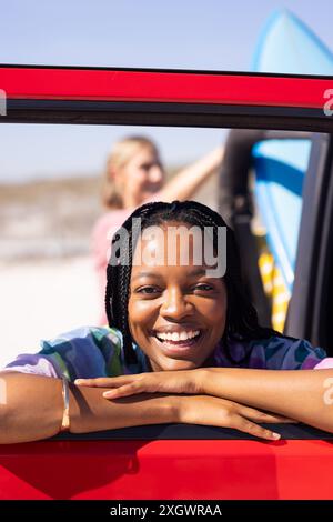 Young African American woman leans out of a car window on a road trip, with copy space. Outdoor setting captures a joyful road trip vibe with surfboar Stock Photo