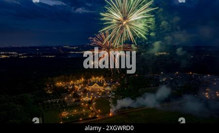 Vibrant Green And Orange Fireworks Burst Over A Lit Estate Amidst Lush Greenery, With Distant City Lights Flickering, Creating A Mesmerizing Nighttime Celebration Scene. Stock Photo