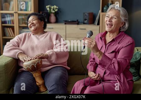 Candid portrait of two cheerful senior women playing music together and singing to microphone at home Stock Photo