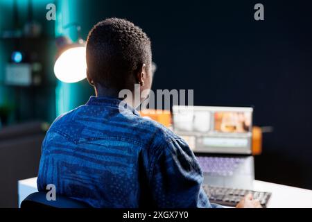 An African American woman edits footage on her laptop at her home office desk, immersed in creative work. Modern studio setup with digital device and software for productive filmmaking. Stock Photo