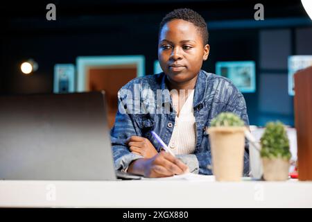 Focused African American woman works from home, browsing the internet, studying online, and multitasking in her modern living room. Female blogger holding pen while looking at laptop. Stock Photo