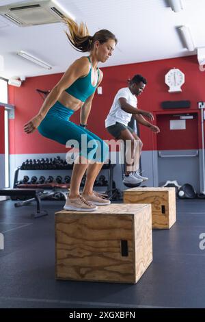 Fit young Caucasian woman and African American man perform box jumps at the gym. Their workout demonstrates strength and agility in a fitness environm Stock Photo