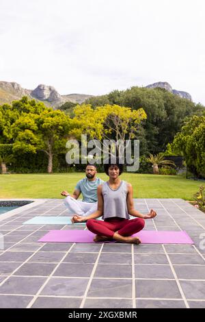 Biracial couple practices yoga outdoors, with copy space. They enjoy a serene session in a tranquil garden setting. Stock Photo