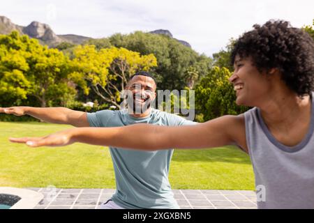 Biracial couple enjoys a fun moment outdoors doing yoga. They're sharing laughter while stretching in yoga poses in a sunny garden. Stock Photo