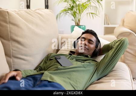 Young biracial man relaxes on a couch at home, with copy space unaltered. He enjoys music with headphones, creating a serene atmosphere. Stock Photo