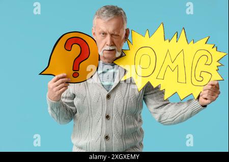 Shocked senior man holding speech bubbles with question mark and word OMG on blue background Stock Photo