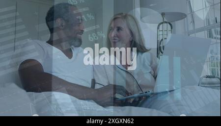 Image of infographic interface moving over happy diverse couple using laptop on bed at home. Digital composite, business, report, chart, love, togethe Stock Photo