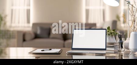 A laptop computer with a white-screen mockup, a clipboard paper, stationery, and decor plants on a table in a cozy living room. home workspace concept Stock Photo