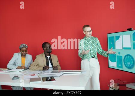 Businesswoman delivering presentation in modern office with red wall and large display. Colleagues attentively listening and taking notes during discussion Stock Photo