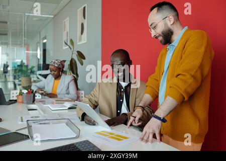 Professionals collaborating on digital project in contemporary office setting. Two men discussing data on digital tablet while woman working independently in background Stock Photo