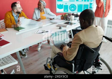 Black man in wheelchair using laptop during team meeting in modern office. Colleagues around table discussing documents, with presentation screen in background Stock Photo