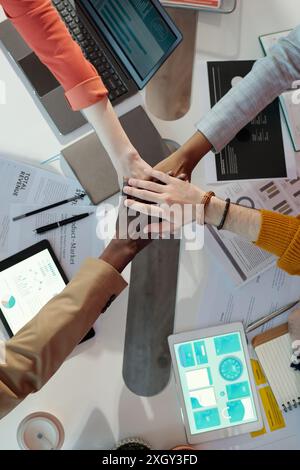 Close-up of team members stacking hands over desk in collaborative office meeting. Diverse hands symbolizing unity and teamwork with digital devices and documents in background Stock Photo