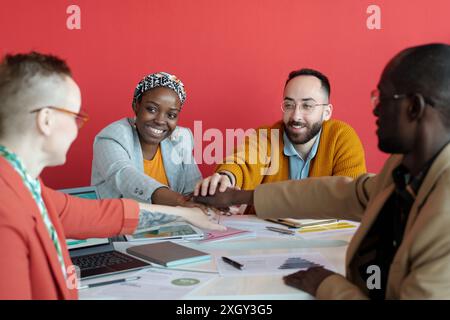 Group of diverse colleagues in office celebrating success by stacking hands together over desk. Smiling professionals demonstrating teamwork and unity in collaborative workspace Stock Photo