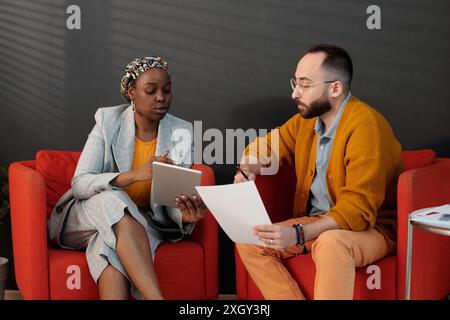 Two colleagues sitting on red chairs, discussing project details, using tablet and document, collaborating and sharing insights in office Stock Photo