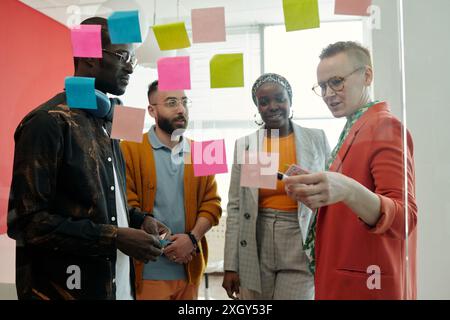 Colleagues gathered around glass wall with colorful sticky notes, collaborating on innovative solutions, discussing and sharing ideas in office Stock Photo