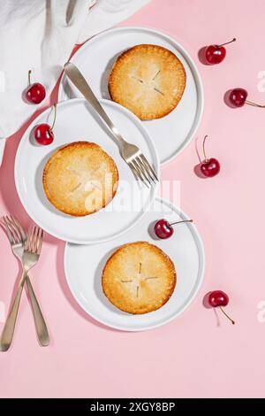 Three cherry pies on white plates with forks, surrounded by fresh cherries on a pink background. Stock Photo
