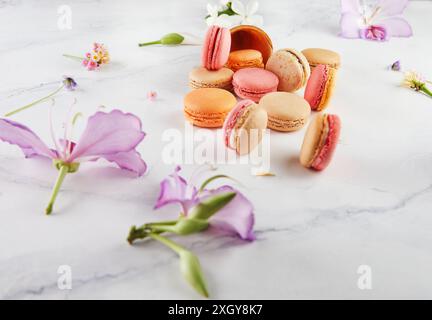 Assorted colorful macarons scattered with an empty ice cream cone and flowers on a white marble background. Stock Photo