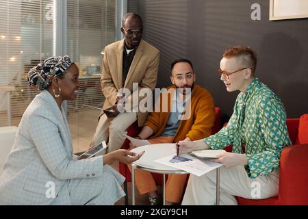 Group of four diverse colleagues sitting in office discussing business statistics and strategies while collaborating on project Stock Photo