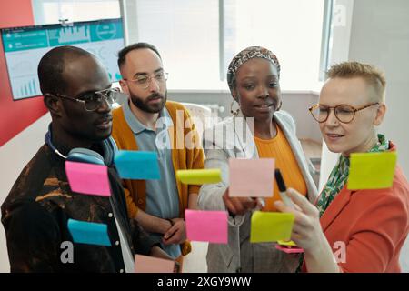 Group of colleagues focused on sticky notes on glass wall, collaboratively discussing strategies and sharing ideas for project in office Stock Photo
