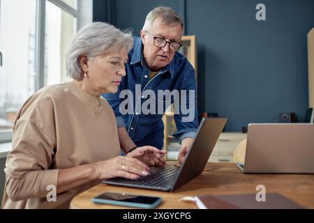Side view portrait of senior woman using laptop in computer class for adults with teacher assisting copy space Stock Photo