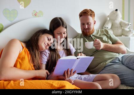 Father and two daughters are reading a book together on a bed, with the father enjoying a cup of tea Stock Photo