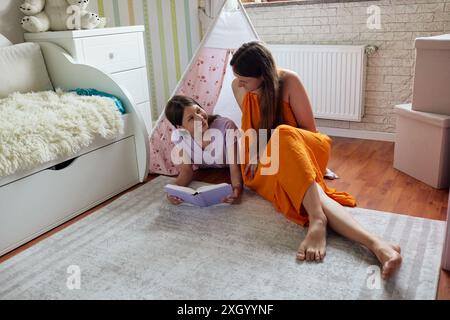 Mother and daughter are reading a book together on the floor of a bedroom, with a white teepee tent nearby Stock Photo