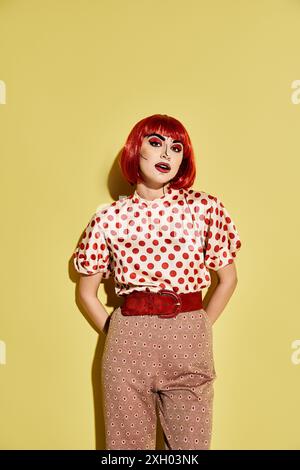 A striking red-haired woman with pop art makeup and a polka dot shirt stands confidently in front of a bright yellow wall. Stock Photo