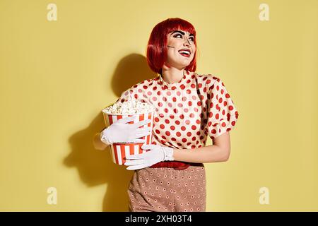 A pretty redhead woman with pop art makeup and white gloves holding a cup of popcorn, dressed in a polka dot blouse. Stock Photo