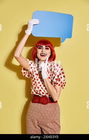 A creative redhead woman in polka dot blouse holding a speech bubble above her head on a yellow background. Stock Photo