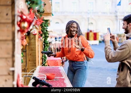 Cheerful African woman plays at a festive shooting gallery in a city Christmas market. Man captures the moment on smartphone, showcasing diverse holid Stock Photo