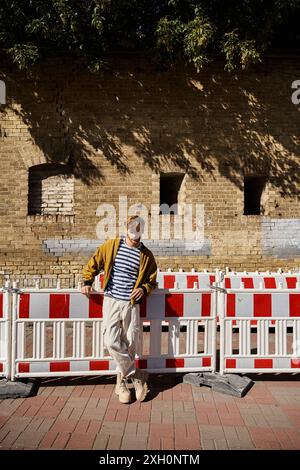 A stylish young man with red hair standing elegantly next to a white and red fence in the city. Stock Photo
