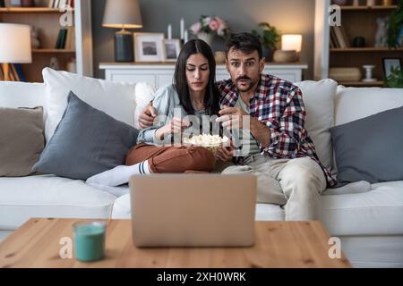 Couple shocked watching horror movie on laptop computer sitting at sofa at home eating popcorn. Fear on their faces, scary film and weekend activities Stock Photo