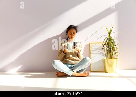 Full body of relaxed young Asian female drinking coffee and browsing social networks on mobile phone while sitting on floor and chilling in light room Stock Photo