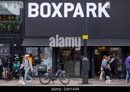 People pass boutique shops at Boxpark in Shoreditch on 3rd July 2024 in London, United Kingdom. Boxpark is a food and retail park made out of refurbished shipping containers. It was founded by Roger Wade, who originally described it as the ‘worlds first pop-up mall’. Boxpark Shoreditch was launched in 2011, and has since pioneered two further Boxparks in Croydon and Wembley. Shoreditch is a fashionable area adjacent to the equally hip neighborhood of Hoxton. Young creatives and trendsetters fill the surroundings with an eclectic scene featuring everything from trendy boutique shops to the ever Stock Photo