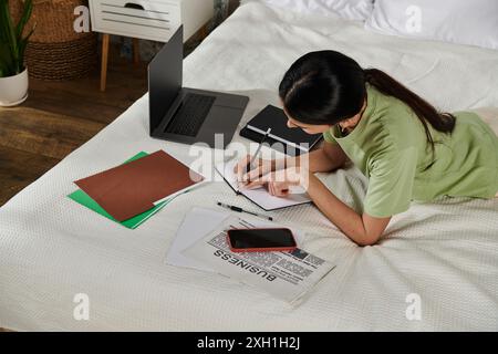 A woman in casual attire sits on a bed, focused on writing in a notebook. Stock Photo