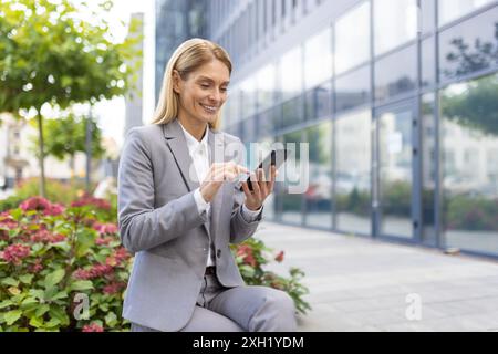 Businesswoman in gray suit using smartphone outdoors at office complex. Professional woman smiling and interacting with technology. Modern urban setting with glass building and greenery. Stock Photo