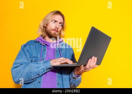 Photo portrait of lovely young male hold netbook confused staring dressed stylish jeans garment isolated on yellow color background Stock Photo