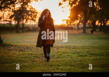 A woman in a brown coat walks through a park, talking on her phone. The sun sets behind her in the distance, casting a warm glow on the scene. Stock Photo