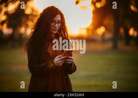A young woman wearing glasses and a coat checks her smartphone in a park at sunset. Stock Photo