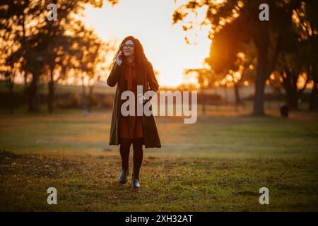 A woman in a coat and glasses walks through a park while speaking on her cell phone. The sun is setting, casting a warm glow over the scene. Stock Photo