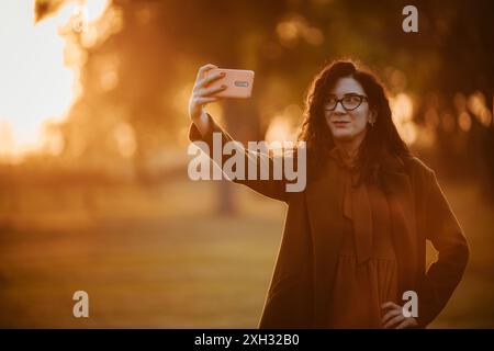 A woman in a brown jacket and glasses takes a selfie with a smartphone in a park, the warm evening sun shining behind her. Stock Photo